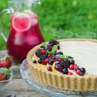 Lemon Curd and Mascarpone Cheesecake with Berries www.thefoodiecorner.gr Photo description: A close look at half the cheesecake, sitting on a glass cake stand on a wooden box in a field. Red berries decorate the one side of the surface cheesecake. A few spearmint leaves are on top of the berries. In the background, a jug of pink lemonade on the box and long green grass behind that.