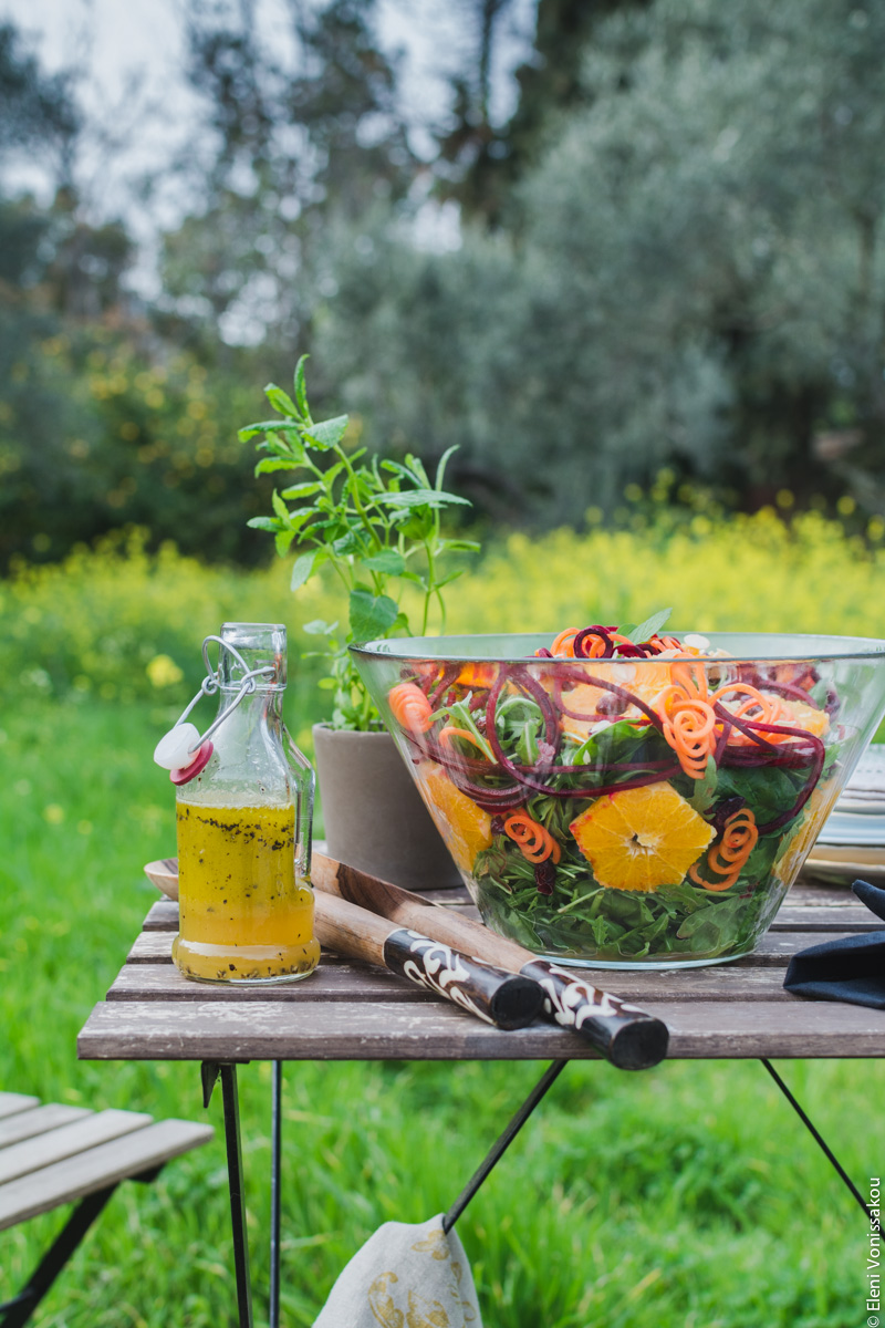 Spinach and Rocket Salad with Orange, Carrot and Raw Beetroot www.thefoodiecorner.gr Photo description: A side view of the table from a little further away. In the background some olive trees, very tall wild yellow flowers and lots of long lush grass.