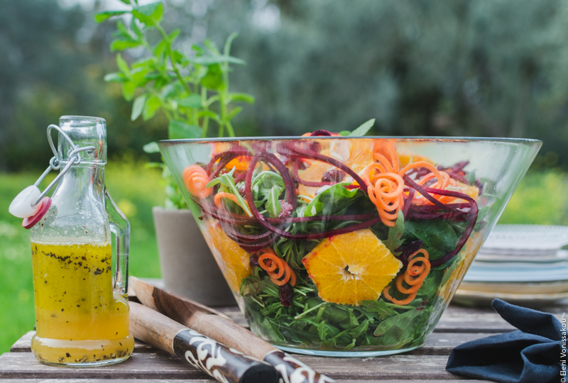 Spinach and Rocket Salad with Orange, Carrot and Raw Beetroot www.thefoodiecorner.gr Photo description: A side view of the large glass bowl of salad, with the contents visible. Flat against the side of the bowl are some star shaped slices of orange and some spirals of carrot. To the left of the bowl is a small bottle of dressing. In the background the small pot of spearmint, some trees and grass.