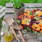 Spinach and Rocket Salad with Orange, Carrot and Raw Beetroot www.thefoodiecorner.gr Photo description: A large glass bowl of colorful salad (half visible) on a small wooden table located outside. On the table are a set of large wooden salad tongs with a pretty pattern on the bottom of the handles, a small bottle with salad dressing inside and a small pot with a spearmint plant. In the background and through the slats of the wooden table, one can see grass. The top of the salad is decorated with star shaped slices of orange, spiralized carrot, spiralized beetroot, almonds and spearmint leaves.