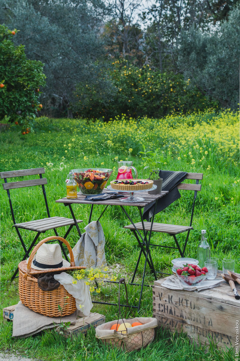 Homemade Pink Lemonade with Beetroot and Ginger www.thefoodiecorner.gr Photo description: A table and two chairs set in a green field with grass and flowers. On the table are a jug of lemonade, a big bowl of salad and a cheesecake. In front of the table on the ground are a picnic basket with a fedora hat on it, a wire basket with some oranges and lemons, and a wooden box with a bowl of strawberries on it. In the background are an orange tree and some olive trees.