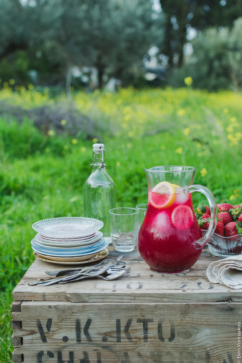 Homemade Pink Lemonade with Beetroot and Ginger www.thefoodiecorner.gr Photo description: The jug of lemonade on the wooden box. To it’s left, a stack of small empty plates and some cutlery right beside it.