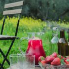 Homemade Pink Lemonade with Beetroot and Ginger www.thefoodiecorner.gr Photo description: A jug of dark pink lemonade with ice cubes and slices of lemon visible through the glass. The jug is on a wooden box set in a lush field with grass and flowers in the background. On the box are some bottles, empty glasses and a bowl of strawberries. To the left there is a wooden chair, half visible.