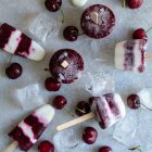 Frozen Cherry Treats – 3 Ways www.thefoodiecorner.gr Photo description: Eight stripy coloured popsicles lying on a stone surface amongst ice cubes and cherries.