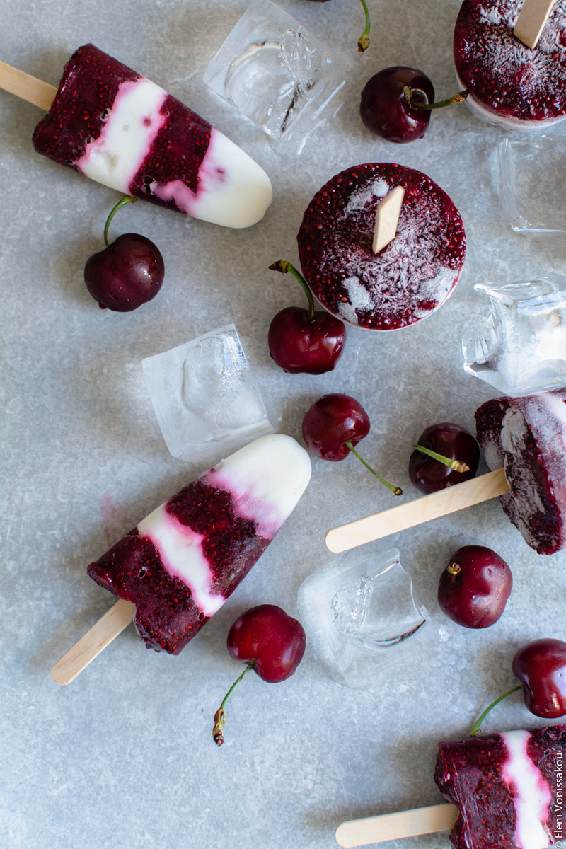 Frozen Cherry Treats – 3 Ways www.thefoodiecorner.gr Photo description: Another, closer view of the stripy coloured popsicles lying on a stone surface amongst ice cubes and cherries.