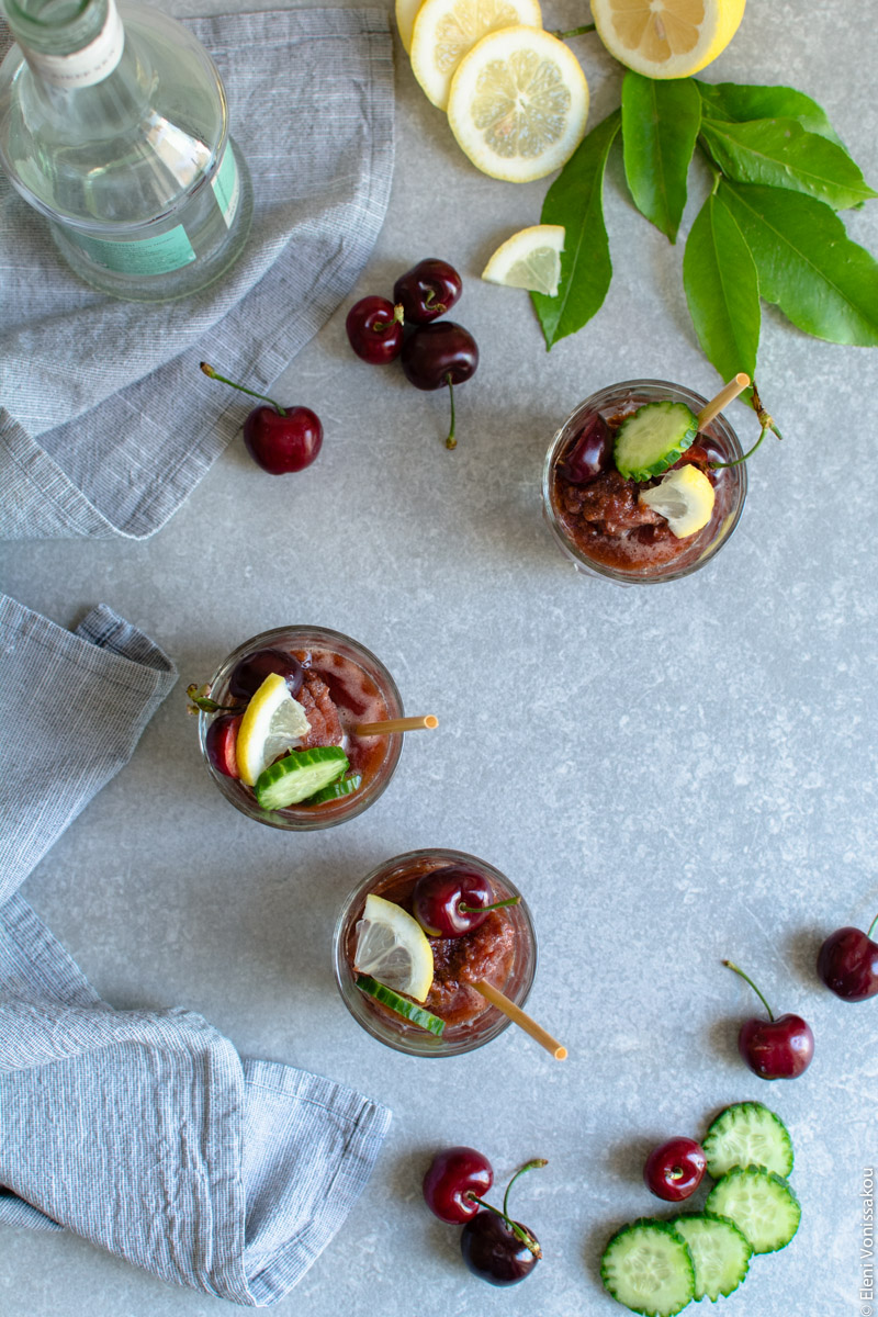 Frozen Cherry Treats – 3 Ways www.thefoodiecorner.gr Photo description: Top view of three short glasses with cherry sorbet inside, on a stone surface. To the top of the photo some lemons with their leaves and a bottle of liqueur, to the left a bunched up tea towel and to the bottom some cucumber and cherry garnishes. The sorbets are decorated with cucumber, cherries and lemon, and there is a wheat straw in each.