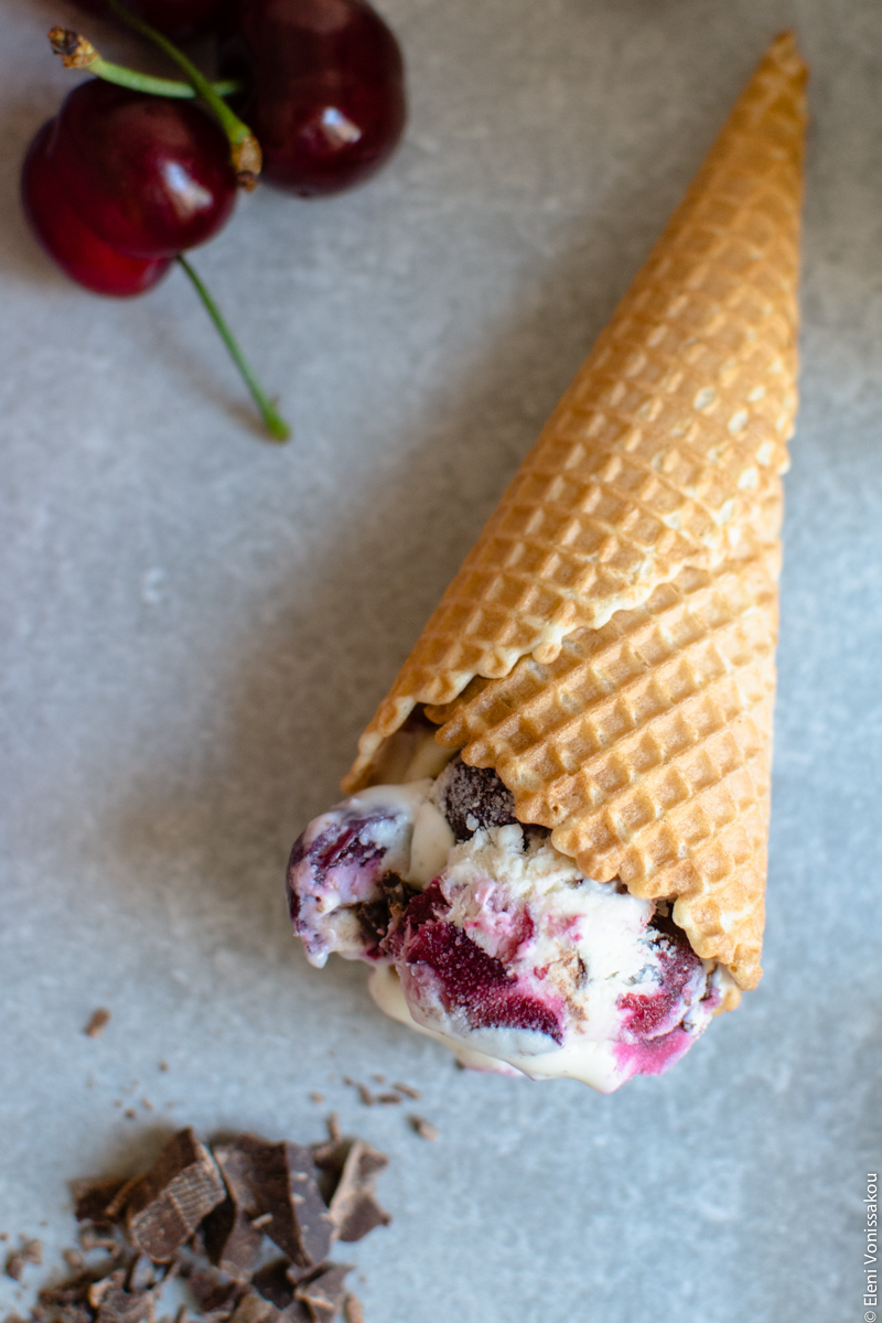 Frozen Cherry Treats – 3 Ways www.thefoodiecorner.gr Photo description: Top view of a wafer cone with ice cream lying on its side on a stone surface. To the top of the photo a few cherries, and to the bottom some chopped dark chocolate.