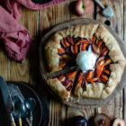 Easy Anthotyro Galette with Spiced Summer Fruit www.thefoodiecorner.gr Photo description: A galette with sliced peaches, apricots and plums, sitting on a round wooden cutting board in the centre of the photo and a little towards the right. One slice has been cut and is slightly pulled back from the galette. In the centre of the galette is a ball of vanilla ice cream. In the bottom left corner of the photo are some stacked plates with cutlery on top. Scattered on the wooden surface of the scene are halved stone fruits. In the top right corner a tiny bowl with honey and a honey wand. To the top left is a linen napkin.