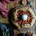 Easy Anthotyro Galette with Spiced Summer Fruit www.thefoodiecorner.gr Photo description: A galette with sliced peaches, apricots and plums, sitting on a round wooden cutting board in the centre of the photo and a little towards the right. One slice has been cut and is slightly pulled back from the galette. In the centre of the galette is a ball of vanilla ice cream. In the bottom left corner of the photo are some stacked plates with cutlery on top. Scattered on the wooden surface of the scene are halved stone fruits. In the top right corner a tiny bowl with honey and a honey wand. To the top left is a linen napkin.