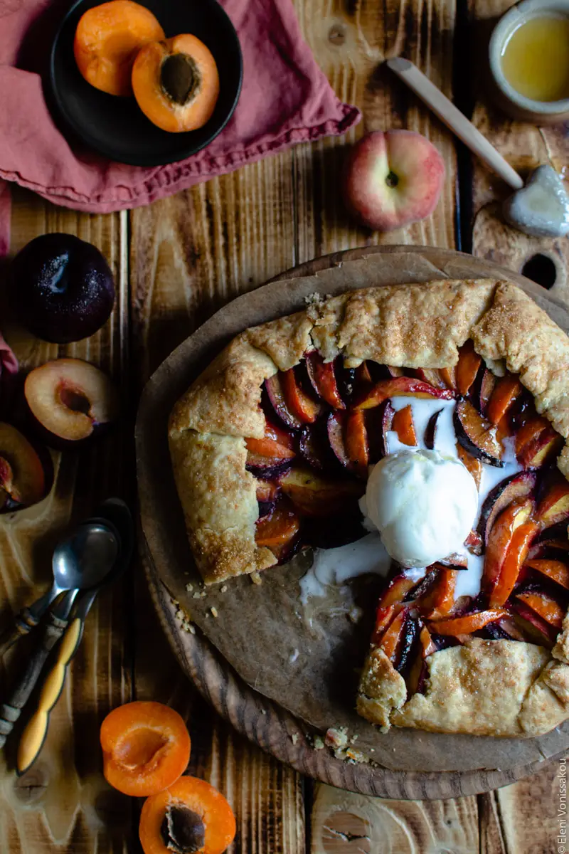 Easy Anthotyro Galette with Spiced Summer Fruit www.thefoodiecorner.gr Photo description: The galette with the melting ice cream on top. Surrounding it are halved apricots and plums and a whole peach. Everything is on an old wooden surface.