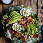 Chicken, Bacon, Avocado and Egg Salad with Gorgonzola Dressing (aka Cobb Salad) www.thefoodiecorner.gr Photo description: A large cobb salad on a platter, set on a wooden surface. To the top some wooden plates barely seen, with a few forks on top. Also to the top a small bowl of creamy dressing. To the bottom of the photo a tea towel made of a jeans-like fabric. The salad itself is decorated with slices of avocado spread out like a fan, sliced chicken breasts, clusters of bacon strips and overlapping slices of egg. Halved cherry tomatoes are dotted here and there.