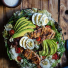 Σαλάτα Κόμπ (Cobb Salad) με Κοτόπουλο, Μπέικον και Ντρέσινγκ Γκοργκοντζόλας www.thefoodiecorner.gr Photo description: A large cobb salad on a platter, set on a wooden surface. To the top some wooden plates barely seen, with a few forks on top. Also to the top a small bowl of creamy dressing. To the bottom of the photo a tea towel made of a jeans-like fabric. The salad itself is decorated with slices of avocado spread out like a fan, sliced chicken breasts, clusters of bacon strips and overlapping slices of egg. Halved cherry tomatoes are dotted here and there.