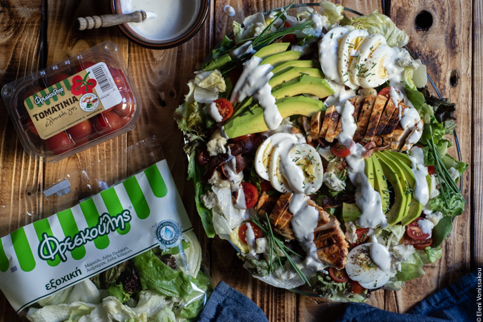 Σαλάτα Κόμπ (Cobb Salad) με Κοτόπουλο, Μπέικον και Ντρέσινγκ Γκοργκοντζόλας www.thefoodiecorner.gr Photo description: The salad with some dressing drizzled over the top in lines. To the left of the platter is a bag of lettuce and a punnet of cherry tomatoes.