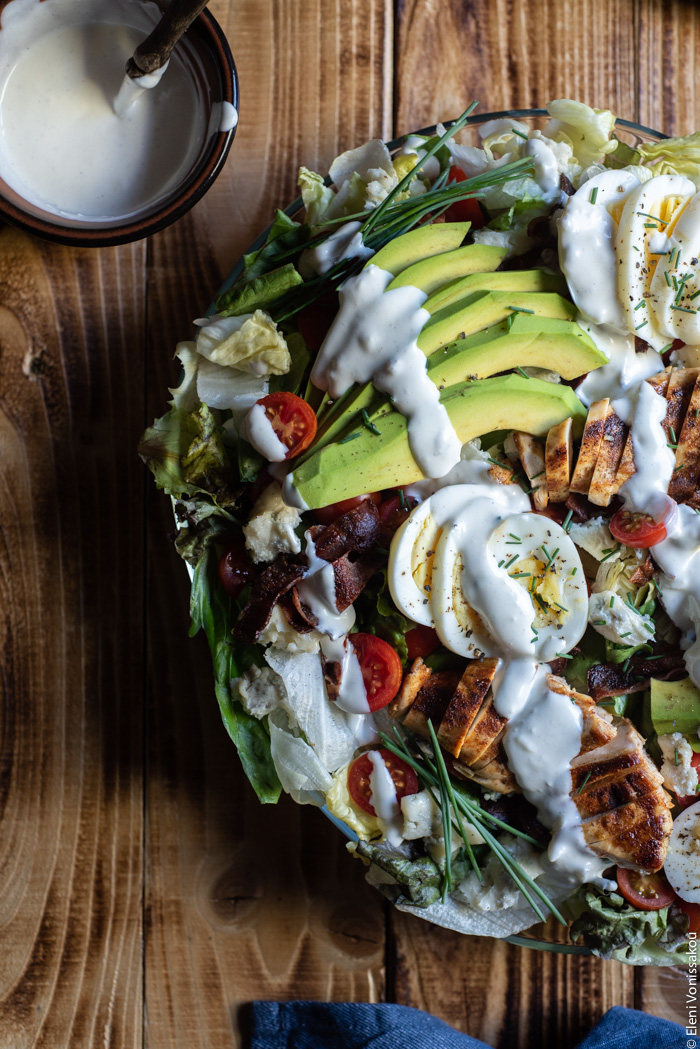 Σαλάτα Κόμπ (Cobb Salad) με Κοτόπουλο, Μπέικον και Ντρέσινγκ Γκοργκοντζόλας www.thefoodiecorner.gr Photo description: A closer view of the cobb salad with the dressing drizzled over it. The salad is half visible. To the top left the bowl of dressing.