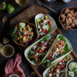 Spicy Salmon Tacos with Manouri Cheese and Peach Avocado Salsa www.thefoodiecorner.gr Photo description: a large wooden tray with tacos laying side by side, open side up, propped up on each side by a bowl of salsa and some lime wedges. Around the tray are some glasses of beer, a linen tea towel and a bowl of flaked salmon.