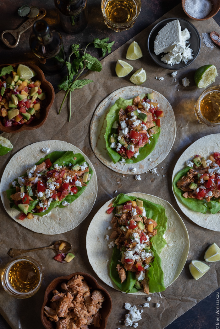 Spicy Salmon Tacos with Manouri Cheese and Peach Avocado Salsa www.thefoodiecorner.gr Photo description: Four tacos laying open on a piece of parchment paper. Around them are some lime wedges, some glasses of beer, a bowl of flaked salmon and a bowl of salsa.