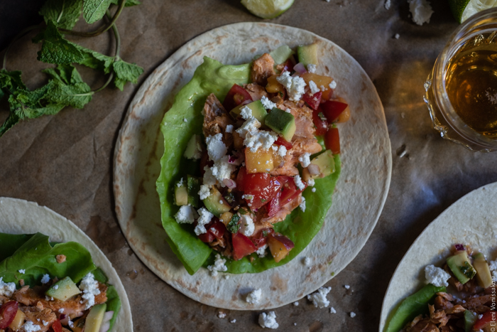 Spicy Salmon Tacos with Manouri Cheese and Peach Avocado Salsa www.thefoodiecorner.gr Photo description: A close up of one of the tacos laying open on parchment paper. Lettuce, salmon flakes, salsa with peach and avocado, and crumbled manouri cheese are all piled on the taco.