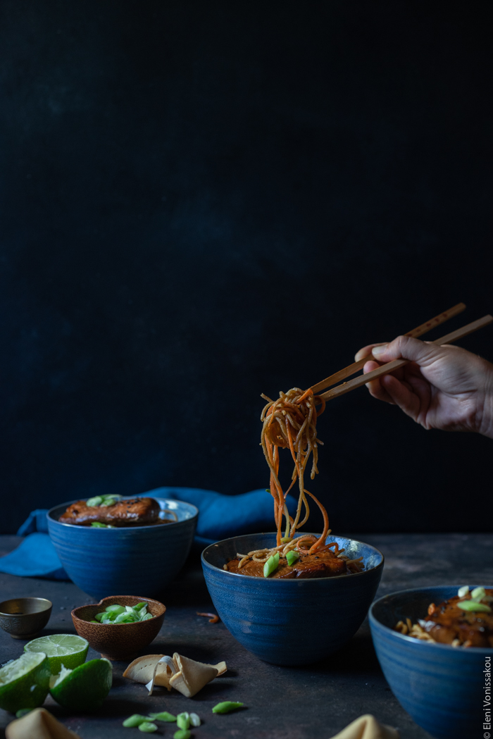 Easy Lo Mein with Salmon, Spaghetti, Veggie Noodles and Peanut Sauce www.thefoodiecorner.gr Photo description: Side view of the bowls of noodles against a dark background. A hand is visible on the right side, holding chop-sticks with a mouthful of noodles hanging down into the bowl.