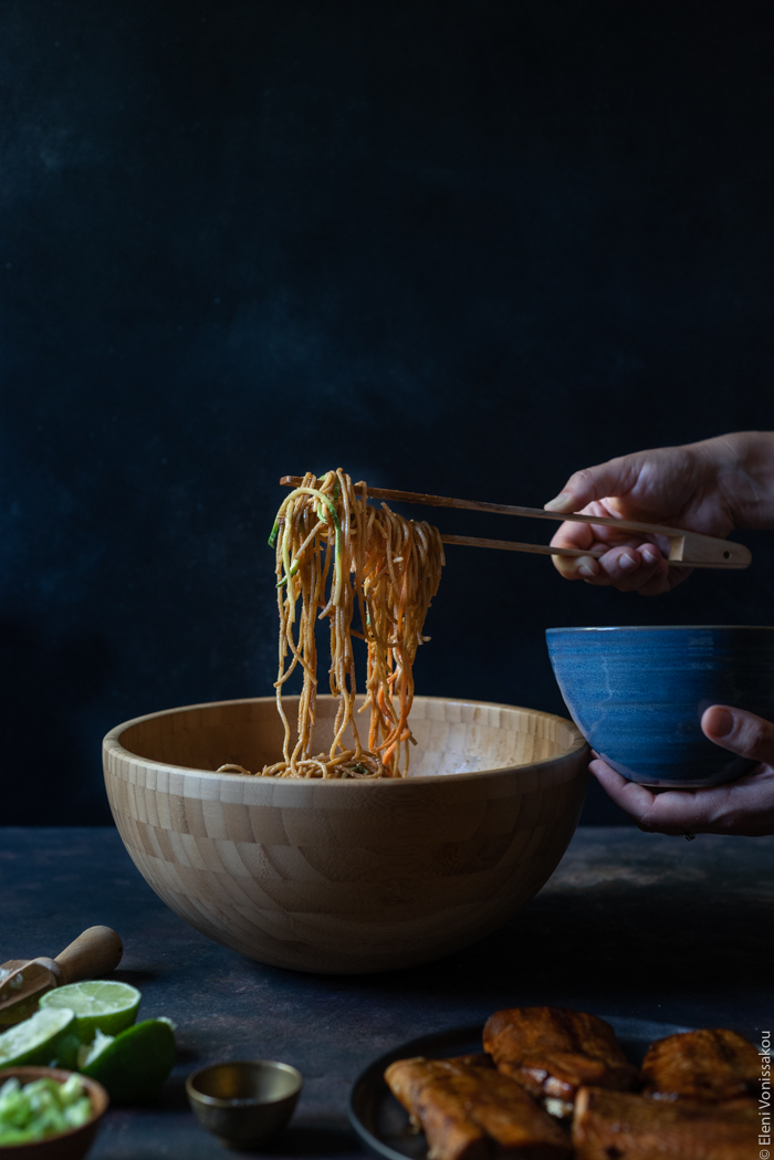 Εύκολο Lo Mein με Σολομό, Σπαγγετίνι Ολικής και Νούντλς Λαχανικών www.thefoodiecorner.gr Photo description: A side view of the big bowl of veggie noodles mixed with whole wheat spaghetti and peanut sauce. On the right a hand just visible with a pair of tongs, holidng up a bunch of noodles from the bowl.