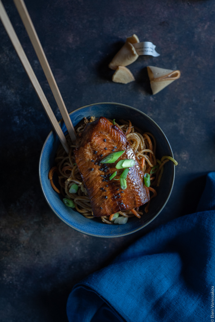 Easy Lo Mein with Salmon, Spaghetti, Veggie Noodles and Peanut Sauce www.thefoodiecorner.gr Photo description: A closer view of one of the bowls with noodles and a salmon fillet on top. The food is topped with some spring onions and black sesame seeds. A pair of chop-sticks are sticking out of the bowl. Right beside it are some fortune cookies and a bunched-up linen napkin.