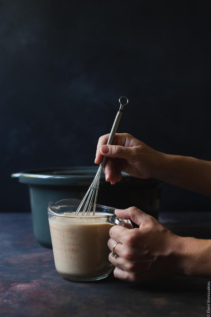 Πουτίγκα Τσουρεκιού στο Slow Cooker με Μπανάνα, Φυστικοβούτυρο και Κομματάκια Σοκολάτας www.thefoodiecorner.gr Photo description: A measuring jug with custard mixture sitting in front of the slow cooker insert. Two hands are visible from the side, one holding the jug the other whisking the mixture.