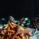 Balsamic Maple Roasted Carrots, Brussels Sprouts and Shallots (Vegan) www.thefoodiecorner.gr Photo description: A side view of a ceramic serving plate filled with roasted vegetables piled high. In the darkened background are some pine cones and ribbons, adding to the festive feel of the image.