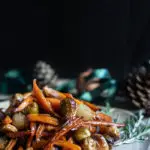 Balsamic Maple Roasted Carrots, Brussels Sprouts and Shallots (Vegan) www.thefoodiecorner.gr Photo description: A side view of a ceramic serving plate filled with roasted vegetables piled high. In the darkened background are some pine cones and ribbons, adding to the festive feel of the image.