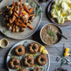 Festive Sweet Potato, Mushroom and Chestnut Mini Nut Loaves www.thefoodiecorner.gr Photo description: A table with large platters of food. One platter has muffin shaped nut loaves, decorated with orange zest and sunflower seeds. The other has roasted Brussels sprouts and carrots. To the top of the photo are a bowl of mushroom gravy and a smaller plate with mashed potatoes. Some orange wedges and chopped chives are scattered on the wooden surface of the table.