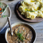 Sweet Wine Mushroom Gravy www.thefoodiecorner.gr Photo description: A bowl of mushroom gravy, some of the sliced mushrooms visible on the surface of the sauce. Some chives decorate the top and a spoon with a wooden handle is inside the gravy. In the background a bowl of mashed potatoes decorated with chives.