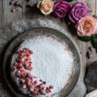 Almond Cardamom Rose Cake with Semolina and Olive Oil www.thefoodiecorner.gr Photo description: A top view of the cake, covered in icing sugar with one side decorated with chopped turkish delight and tiny dried rosebuds. The ceramic dish the cake is on, is sitting on wooly material. Around it, on the bare wooden surface are two vintage cake servers, some roses, and a small bowl of dried rosebuds.