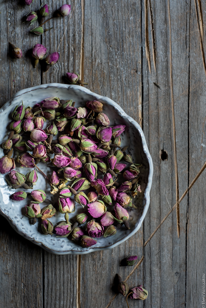 Almond Cardamom Rose Cake with Semolina and Olive Oil www.thefoodiecorner.gr Photo description: a small ceramic bowl full of small dried rosebuds. Some rosebuds are also scatterd around the bowl on the distressed wooden surface.