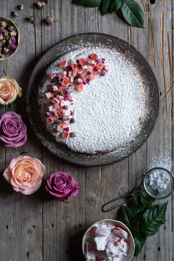 Almond Cardamom Rose Cake with Semolina and Olive Oil www.thefoodiecorner.gr Photo description: The cake on the ceramic plate, some cut roses to its left. To the bottom right of the photo are a bowl of turkish delight, some rose leaves and a small sieve with icing sugar in it. 