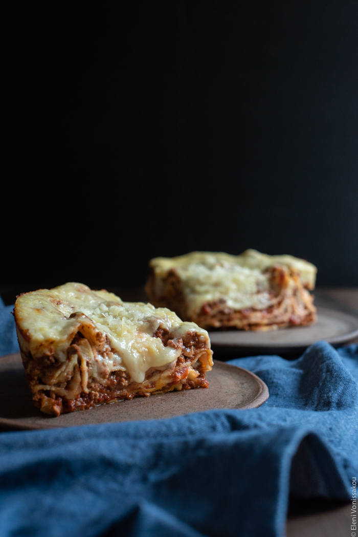 Slow Cooker Lasagna with Meat Sauce and Béchamel www.thefoodiecorner.gr Photo description: A side view of two pieces of lasagna, one of them with a bit of melted cheese oozing down the side. The food is on ceramic plates sitting on a wooden surface, a dark background behind them.