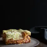Slow Cooker Lasagna with Meat Sauce and Béchamel www.thefoodiecorner.gr Photo description: A side view of a delicious piece of lasagna, the meat and béchamel visible between the layers. The food is on a ceramic plate set against a dark background.