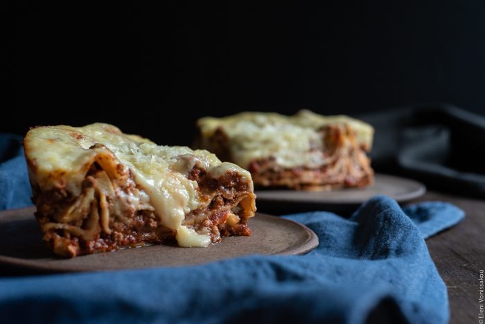 Slow Cooker Lasagna with Meat Sauce and Béchamel www.thefoodiecorner.gr Photo description: A side view of two pieces of lasagna, one of them with a bit of melted cheese oozing down the side. In front of the plates is a blue denim style napkin.