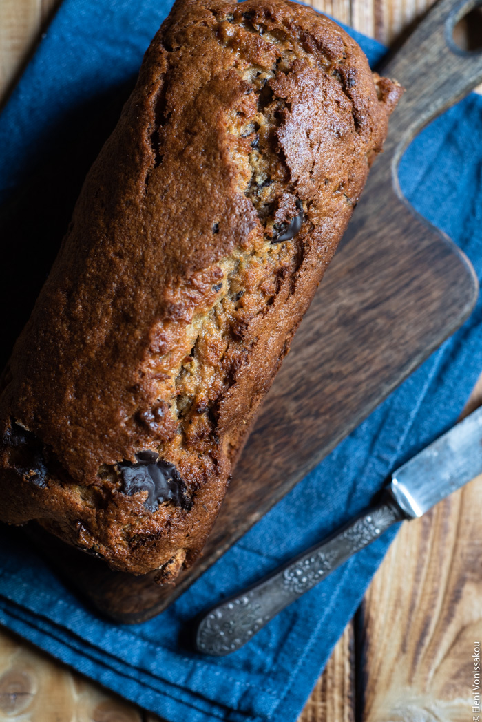 Chocolate Chip Emmer Flour Banana Bread with Tahini www.thefoodiecorner.gr Photo description: A whole banana bread loaf on a wooden chopping board. The board is sitting on a folded tea towel. Next to the loaf is a vintage knife.