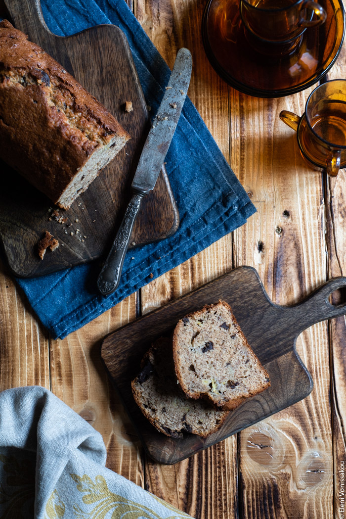 Chocolate Chip Emmer Flour Banana Bread with Tahini www.thefoodiecorner.gr Photo description: A view from higher up. Banana bread on a wooden board, next to it a vintage knife. A couple of pieces cut from the loaf are lying on a smaller wooden board. In the top right corner some small glass coffee cups stacked one on top of the other. In the bottom left corner a bunched up tea towel.