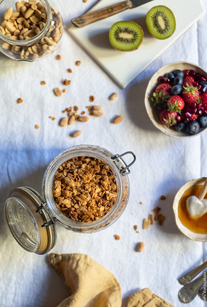 Slow Cooker Peanut Butter Granola with Pure Sesame Oil and Honey www.thefoodiecorner.gr Photo description: An overhead view of a mason jar with granola inside. In the top left corner a jar with peanuts, next to it a small wooden chopping board with a halved kiwi. Further down a small bowl of strawberries and blueberries. At the bottom of the photo a bunched up linen napkin.