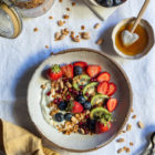 Slow Cooker Peanut Butter Granola with Pure Sesame Oil and Honey www.thefoodiecorner.gr Photo description: An overhead view of a bowl of granola with yogurt, kiwi fruit, strawberries and blueberries, on white linen. In the top left corner an open mason jar with granola, next to it a small bowl with strawberries and blueberries, just below that a smaller bowl with honey and a honey wand lying inside. In the bottom right corner some spoons barely visible. In the bottom left corner a bunched up linen napkin.