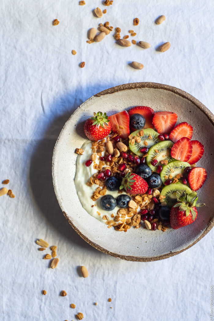 Slow Cooker Peanut Butter Granola with Pure Sesame Oil and Honey www.thefoodiecorner.gr Photo description: An overhead view of a pretty ceramic bowl with granola, yogurt, kiwi fruit, strawberries and blueberries on top. The bowl is on a linen tablecloth and some granola is scattered around it.