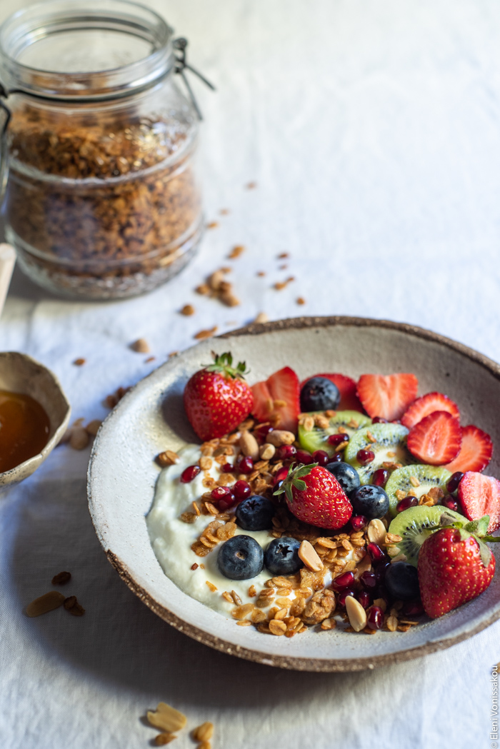 Slow Cooker Peanut Butter Granola with Pure Sesame Oil and Honey www.thefoodiecorner.gr Photo description: A ¾ view of a bowl of granola, yogurt, kiwi fruit, strawberries and blueberries, on white linen. In the background a mason jar with granola inside.