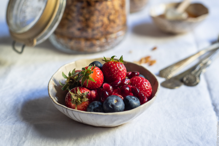 Slow Cooker Peanut Butter Granola with Pure Sesame Oil and Honey www.thefoodiecorner.gr Photo description: A close up view of a small bowl with strawberries and blueberries. A mason jar with granola in the background and a couple of spoons to the right of the photo.