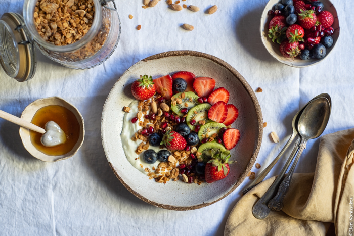 Slow Cooker Peanut Butter Granola with Pure Sesame Oil and Honey www.thefoodiecorner.gr Photo description: An overhead view of a bowl of granola with yogurt, kiwi fruit, strawberries and blueberries, on white linen. In the top left corner an open mason jar with granola, in the top right a small bowl with strawberries and blueberries. In the bottom right corner a bunched up linen napkin and some spoons. To the left of the granola bowl is a small ceramic bowl with honey and a honey wand lying in it.