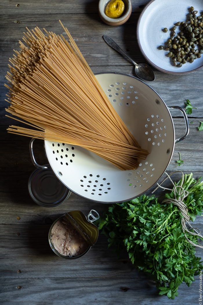 Quick and Easy Smoked Tuna Spaghetti with Parsley and Capers www.thefoodiecorner.gr Photo description: Ingredient shot on a wooden surface. To the top right is a small ceramic plate with capers, next to it a tiny ceramic bowl with English mustard. In the centre of the photo is a large colander with a some uncooked whole wheat spaghetti inside. To the bottom of the colander is a bunch of parsley tied with string and two tins, one of them half opened to show tuna inside.