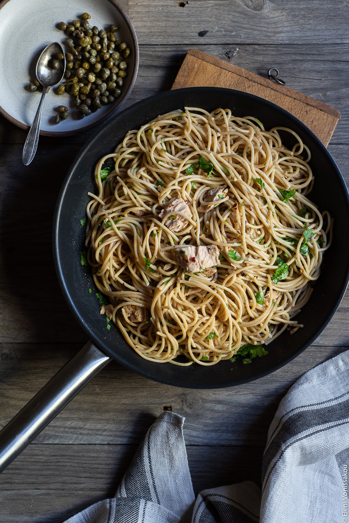 Quick and Easy Smoked Tuna Spaghetti with Parsley and Capers www.thefoodiecorner.gr Photo description: In the centre of the photo is a frying pan full of prepared smoked tuna pasta sitting on a chopping board. Lying beside the pasta in the pan is a wooden cooking utensil. To the bottom of the pan is a bunched up linen tea towel and to the top left is a plate of capers.