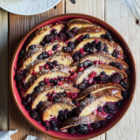 Lemon Berry Tsoureki (Greek Sweet Bread) Pudding www.thefoodiecorner.gr Photo description: A round baking dish full of baked tsoureki pudding. Between the slices are some berries and everything is covered in a sprinkling of icing sugar. To the top of the photo are a small jar of fruit sauce and a small sieve with icing sugar in. To the bottom of the photo a bunched up napkin.