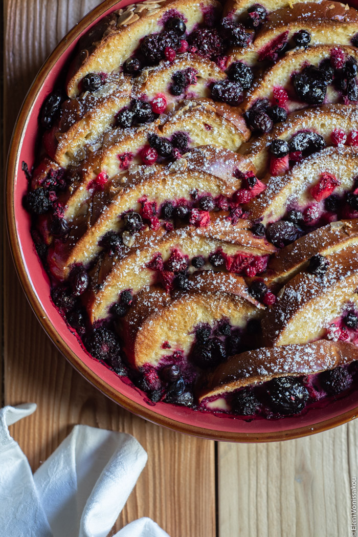 Lemon Berry Tsoureki (Greek Sweet Bread) Pudding www.thefoodiecorner.gr Photo description: A close view of a round baking dish full of baked tsoureki pudding. Between the slices are some berries and everything is covered in a sprinkling of icing sugar.