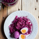 Quick Pickled Eggs and Red Cabbage www.thefoodiecorner.gr Photo description: A plate with a halved egg and some pickled cabbage on it. The egg’s outside surface has a lilac hue to it from the cabbage. To the top of the photo is an open jar with purple pickling liquid visible inside.