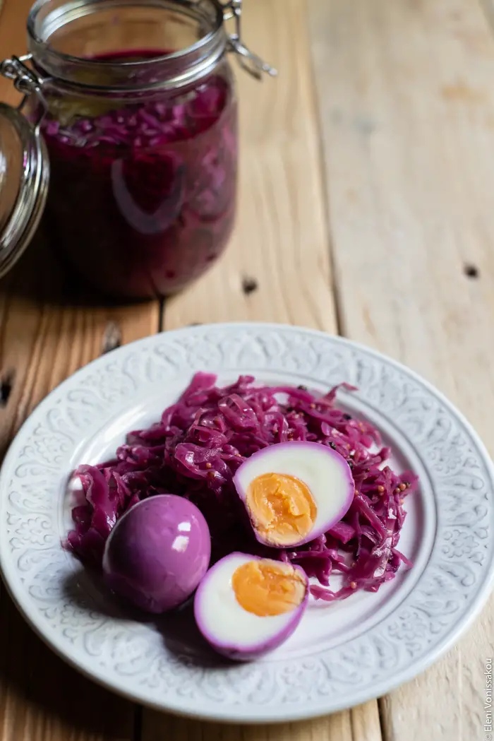 Quick Pickled Eggs and Red Cabbage www.thefoodiecorner.gr Photo description: A ¾ view of a plate with one whole and one halved pickled egg and some pickled red cabbage. In the background a jar with pickling juice inside.