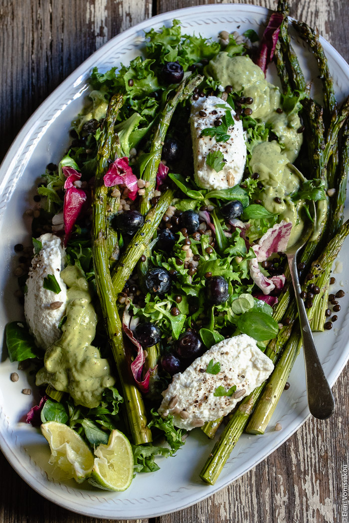 Green Salad with Buckwheat, Beluga Lentils, Roasted Asparagus and Avocado Dressing www.thefoodiecorner.gr Photo description: A close view of a salad on a platter, consisting of green salad leaves, asparagus, beluga lentils, buckwheat and blueberries, tossed together. Some soft white cheese and some salad dressing on a spoon are also visible, as well as some lime wedges. 